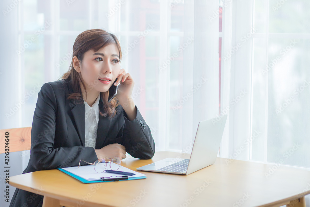 The office worker, a beautiful Asian woman with a smiling and happy face, is sitting talking on the phone at her desk. On the desk there is a laptop and documents. The office worker, a beautiful Asian woman with a smiling and happy face, is sitting talking on the phone at her desk. On the desk there is a laptop and documents.