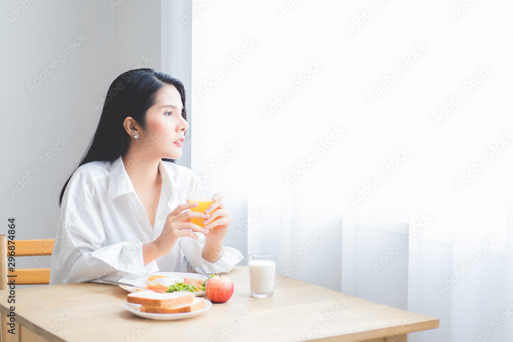 A beautiful Asian woman, a tan, wearing a long-sleeved white shirt and shorts, and on her hand holding a fork and a knife, is eating breakfast in a bedroom at a hotel in Thailand