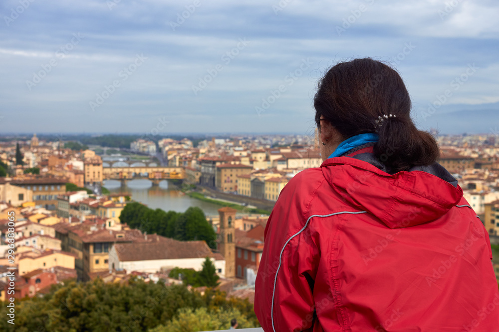 Naklejka premium Woman tourist in a red jacket looks at the panorama of historical center of Florence in Italy from Piazzale Michelangelo. Back view. Arno river, the Duomo and many other churches and buildings