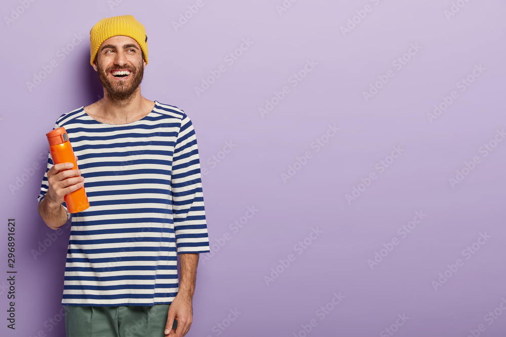 Cheerful unshaven millennial man holds flask with hot beverage, wears yellow hat and striped jumper, being in good mood, stands against purple background, copy space for your promotional content