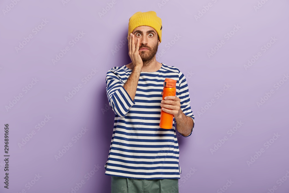 Astonished tourist has coffee break, holds flask with beverage, touches cheek, wears casual clothes, cannot believe in something unbelievable, gets unexpected rumors, isolated over purple background