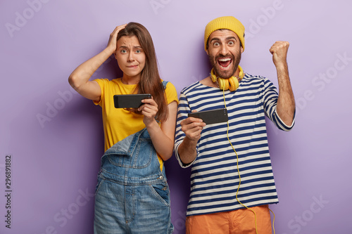 Young woman and man play video games on smartphones, being addicted to modern technologies, guy shows fist bump, rejoices victory, wears striped jumper and headphones, worried female stands near