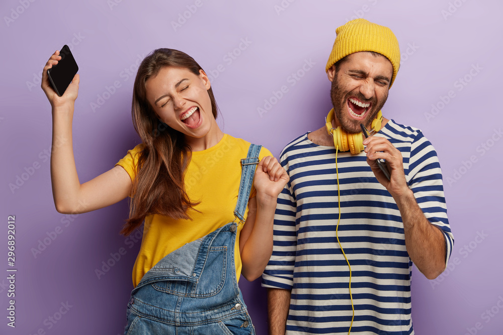 Candid shot of overjoyed energized boyfriend and girlfriend listen music via cell phone, dance and sing loudly, express positive emotions, stand next to each other, raise arms and move actively Candid shot of overjoyed energized boyfriend and girlfriend listen music via cell phone, dance and sing loudly, express positive emotions, stand next to each other, raise arms and move actively