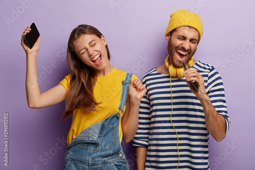 Candid shot of overjoyed energized boyfriend and girlfriend listen music via cell phone, dance and sing loudly, express positive emotions, stand next to each other, raise arms and move actively