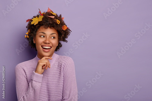 Charismatic lovely woman touches jawline, looks away with happy expression, pleased to be in friendly outgoing company, has autumn leaves on hair, expresses positive emotions, dressed casually