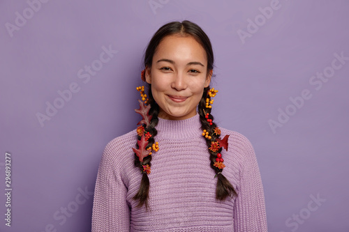 Canvas-taulu Glad natural Asian woman celebrates fall, finds inspiration in nature, has two plaits decorated with red autumn leaves, berries and flowers, wears purple sweater, isolated on violet wall