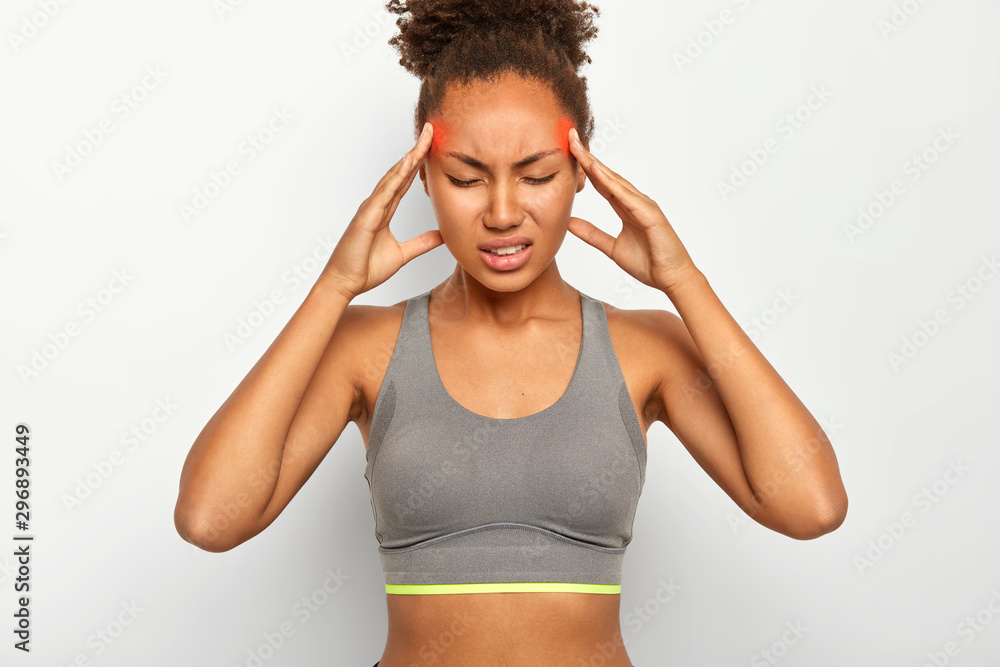 Fatigue overworked Afro American woman massages temples, feels strong headache, closes eyes with pain, suffers from migraine after workout, dressed in sport bra, clenches teeth, isolated on white wall Fatigue overworked Afro American woman massages temples, feels strong headache, closes eyes with pain, suffers from migraine after workout, dressed in sport bra, clenches teeth, isolated on white wall