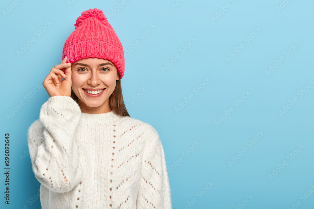 Good looking brunette young woman keeps hand near face, smiles pleasantly, wears hat and white knitted sweater, enjoys pleasant moment, expresses good emotions, poses against blue studio wall Good looking brunette young woman keeps hand near face, smiles pleasantly, wears hat and white knitted sweater, enjoys pleasant moment, expresses good emotions, poses against blue studio wall
