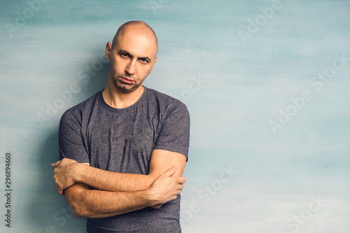Fototapeta A young bald man in a gray T-shirt on a blue background looking angrily at the c