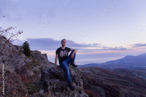 Young attractive man located with on the top of mountains against the backdrop of a purple sunset,