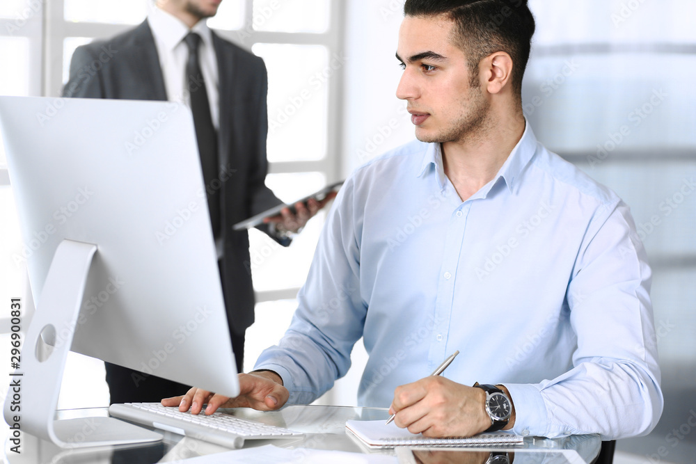 Businessman working with computer in modern office, male colleague at background . Headshot of arab male entrepreneur or manager at workplace. Business concept, teamwork and multi ethnic Businessman working with computer in modern office, male colleague at background . Headshot of arab male entrepreneur or manager at workplace. Business concept, teamwork and multi ethnic