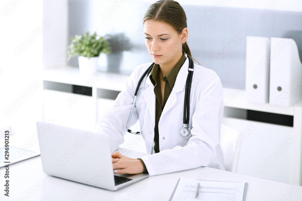 Doctor typing on laptop computer while sitting at the desk in hospital office. Physician woman at work. Data in medicine and healthcare Doctor typing on laptop computer while sitting at the desk in hospital office. Physician woman at work. Data in medicine and healthcare