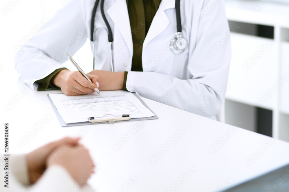 Woman doctor and patient sitting and talking at medical examination at hospital office, close-up. Therapist filling up medication history records. Medicine and healthcare concept Woman doctor and patient sitting and talking at medical examination at hospital office, close-up. Therapist filling up medication history records. Medicine and healthcare concept