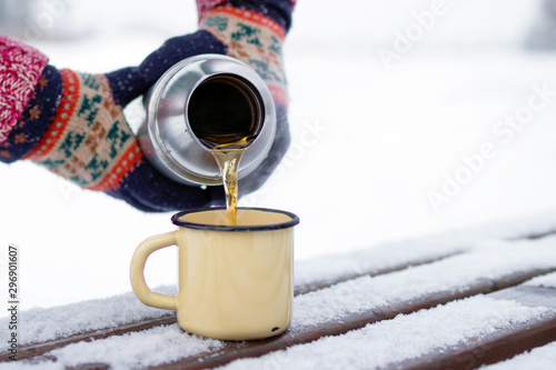 Woman is pouring tea from thermos into cup on a walk in winter park.