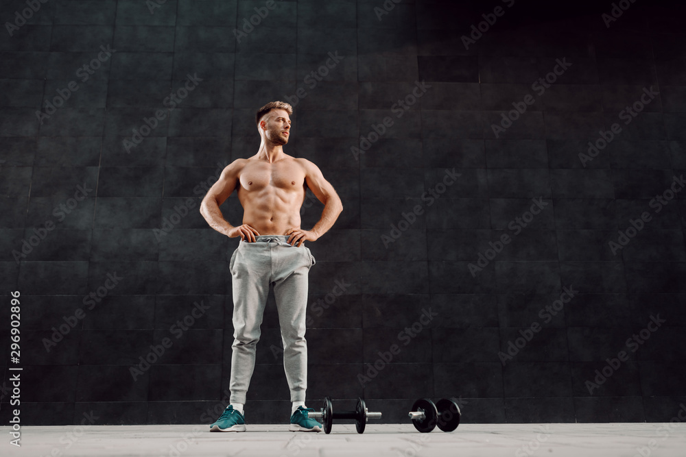 Full length of handsome shirtless muscular caucasian man standing with hands on hips in front of dark background and looking away. Next to his feet are dumbbells.