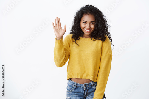 Shy lovely african-american girl in yellow sweater, curly hairstyle, stooping as raising hand, waving hello, hi gesture, smiling, modest woman introduce herself in front audience, white background