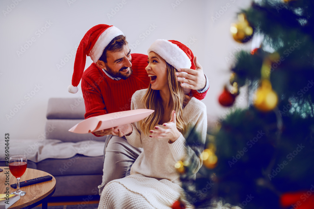 Fototapeta premium Amazed cute Caucasian blonde woman sitting on sofa in living room and receiving gift from her boyfriend. Both having santa hats on heads. In foreground is christmas tree. Living room interior.