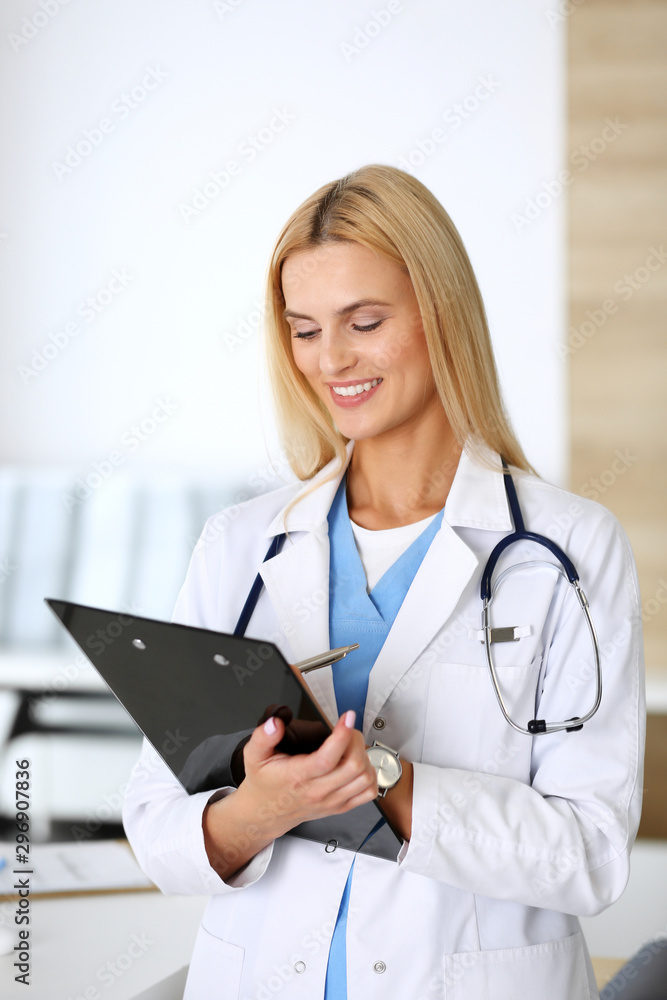 Doctor woman at work in hospital excited and happy of her profession. Blonde physician controls medication history records and exam results while using clipboard. Medicine and healthcare concept Doctor woman at work in hospital excited and happy of her profession. Blonde physician controls medication history records and exam results while using clipboard. Medicine and healthcare concept