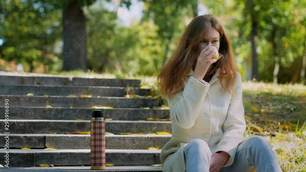 Young girl holding in hands and tasting cup of hot tea on stairs in outdoors autumn park, beautiful woman enjoy drinking cup of coffee thermos, lifestyle relax recreation weekend meditation concept Young girl holding in hands and tasting cup of hot tea on stairs in outdoors autumn park, beautiful woman enjoy drinking cup of coffee thermos, lifestyle relax recreation weekend meditation concept