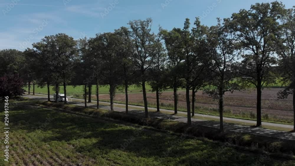 Aerial approach and pan showing countryside with a lane of trees along a country road with backlight following a white camper passing by Aerial approach and pan showing countryside with a lane of trees along a country road with backlight following a white camper passing by