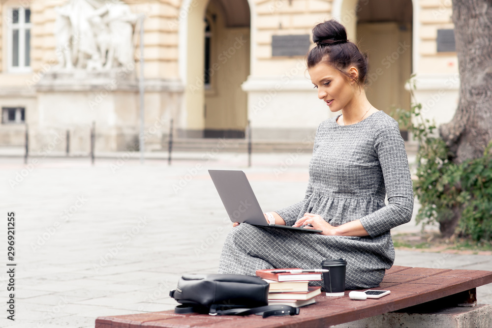Student girl is working on laptop on bench near university building. Female student works on laptop on bench outdoors. Student girl is working on laptop on bench near university building. Female student works on laptop on bench outdoors.