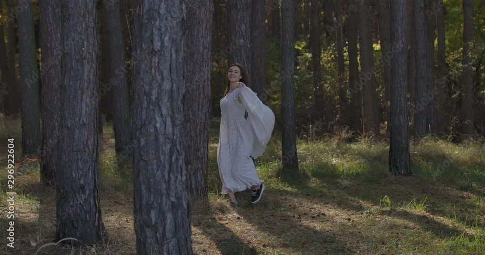Happy Caucasian girl running between the trees in the summer forest and smiling. Mysterious independent woman in light white dress having fun outdoors. Happy Caucasian girl running between the trees in the summer forest and smiling. Mysterious independent woman in light white dress having fun outdoors.