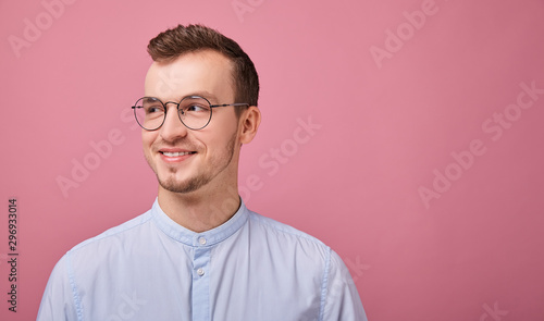 A young man in a sky blue shirt and computer glasses stands on a pink background. Student, businessman, employee of the year. Looking straight with calm.