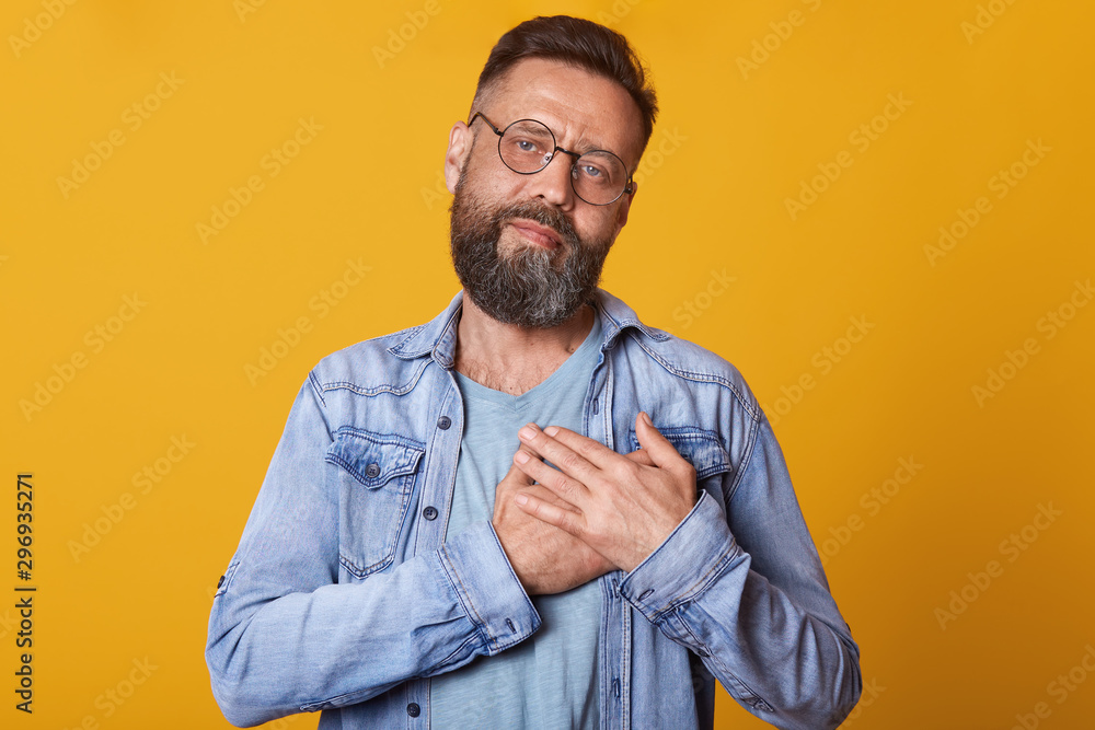 Image of pleased bearded man with rounded optical eyewear, keeps hands on chest, dresses fashionable denim jacket, posing isolated over yellow studio background, expresses his love and grateful. Image of pleased bearded man with rounded optical eyewear, keeps hands on chest, dresses fashionable denim jacket, posing isolated over yellow studio background, expresses his love and grateful.