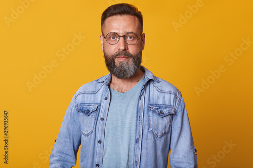Photo of attractive middle aged man with serious and calm facial expression, having beard, dresses casual denim jacket and eyewear, posing isolated over yelow studio wall. People emotions concept.