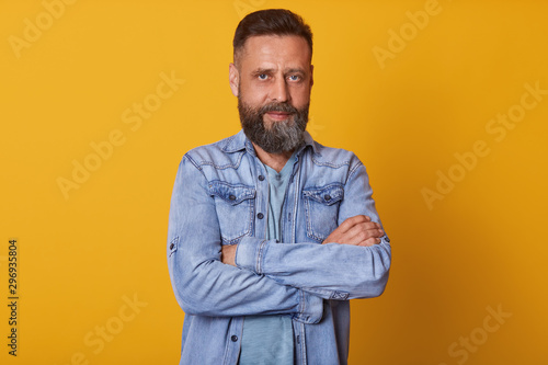 Representative, personable man with beard looking directly at camera, stands with arms folded, isolated over yellow background, dresses stylish denim jacket, having seriousfacial expression.