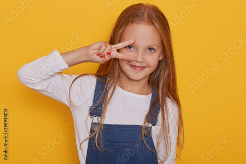 Close up portrait of nice attractive lovely cheerful straight haired blonde girl showing v sign near her eye isolated over bright vivid yellow background, child dresses white shirt. Dody language.