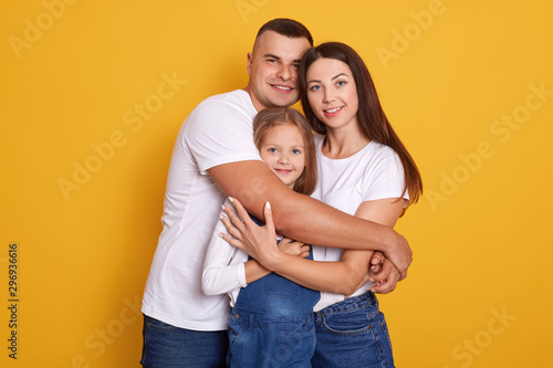 Horizontal shot of beautiful young family hugging, looking directly at camera and smiling while standing against yellow studio wall, people dress casually, expressing happyness. Relationship concept