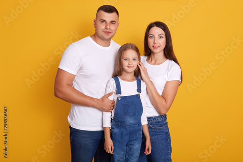 Image of happy Caucasian family pose smiling isolated over yellow background, parents with cute little daughter enjoy spending together, dress casual outfits, looking at camera. Happyness concept.