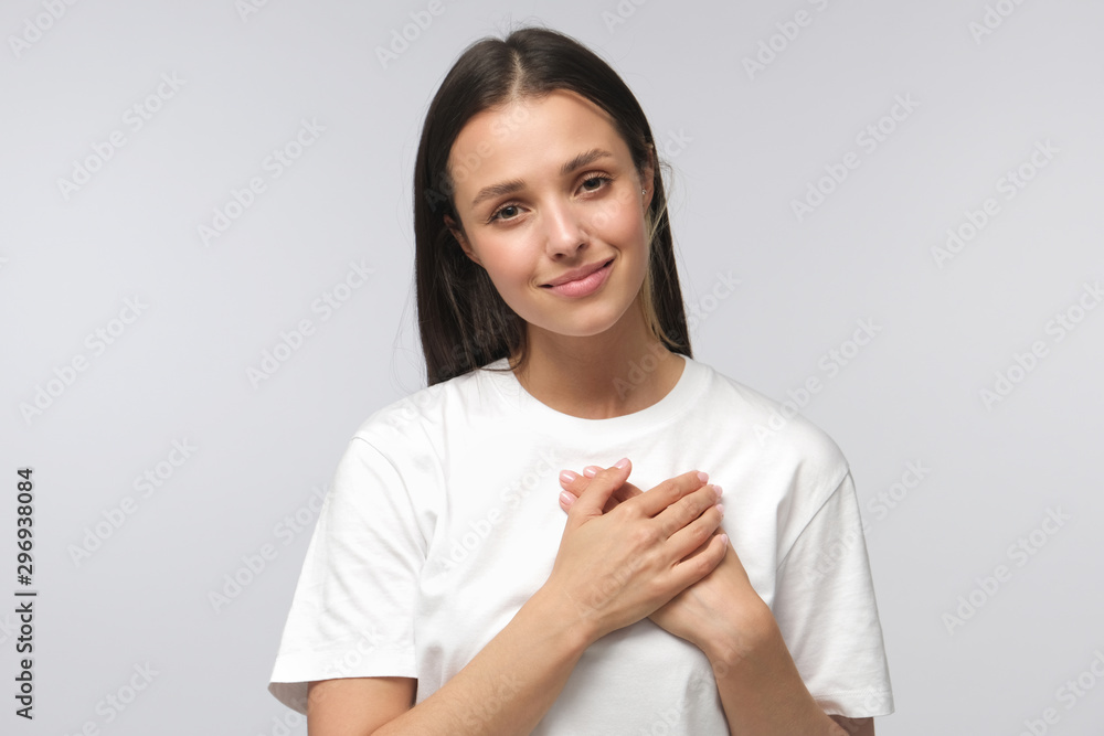 Portrait of smiling positive young woman keeps hands on chest, expresses sympathy. Kind hearted friendly nice female shows kindness, wearing blank white t-shirt Portrait of smiling positive young woman keeps hands on chest, expresses sympathy. Kind hearted friendly nice female shows kindness, wearing blank white t-shirt