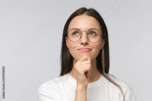 Portrait of young woman with dreamy cheerful expression, thinking, wearing eyeglasses, isolated on gray background
