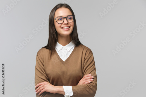 Studio portrait of smiling attractive female in brown sweater standing with crossed arms and looking aside, isolated on gray background