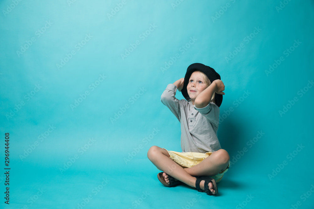 A boy on a blue background sits in a black hat and looks at the camera. The boy is wearing a grey t-shirt. Happy