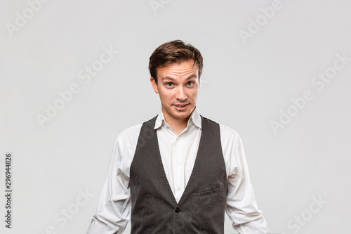 Handsome man in a white shirt and grey vest expressing emotion of surprise stending isolated over light grey background.