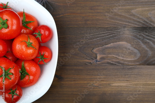 red tomatoes with green tails of different sizes and water drops in a dish on a dark wooden surface with copy space