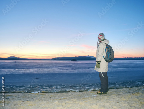 Woman in warm long jacket stay on sandy beach at frozen lake or river. Winter chilly morning with daybreak at hilly horizon.
