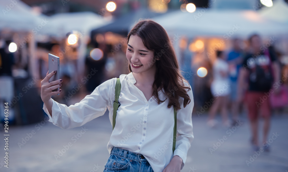 Fototapeta premium Happy traveller young woman using mobile phone to taking selfie at night street market with light bokeh background in Bangkok Thailand.