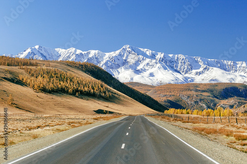 Road To Mountains. Landscape with empty asphalt road through autumn forest, blue sky, high rocks.