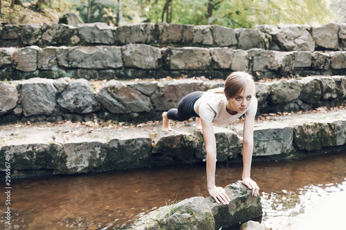 Sports girl stands in a pose between two stones on river. Slender woman doing yoga