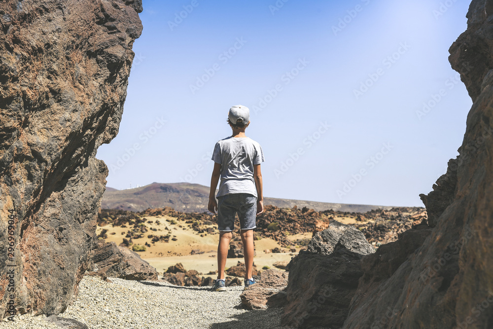 Back view of a teen, standing between lava rocks, looking at the horizon. Trendy boy looks enthusiastically at the view of the Teide National Park. Freedom, travel, tourist and outdoors concept.
