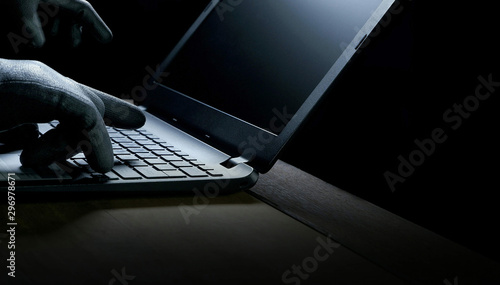 Hacker hands in black gloves using laptop computer on wooden table in dark night background, hacking and internet cyber crime concept
