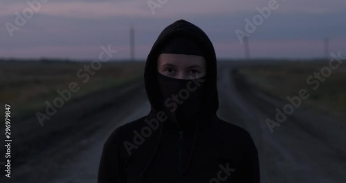 Girl looks into space in front of herself, black balaclava and hood are put on her head, she is standing on country road alone, girl takes off her mask with sharp movement, looks at camera with call