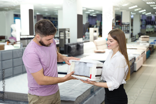Young saleswoman showing sample of modern orthopedic mattress to customer in store