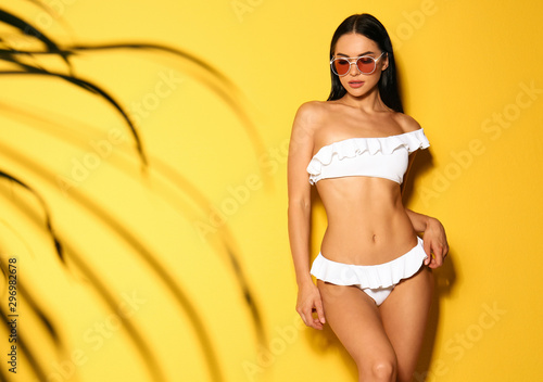Obraz na płótnie Beautiful young woman in white bikini with sunglasses on yellow background