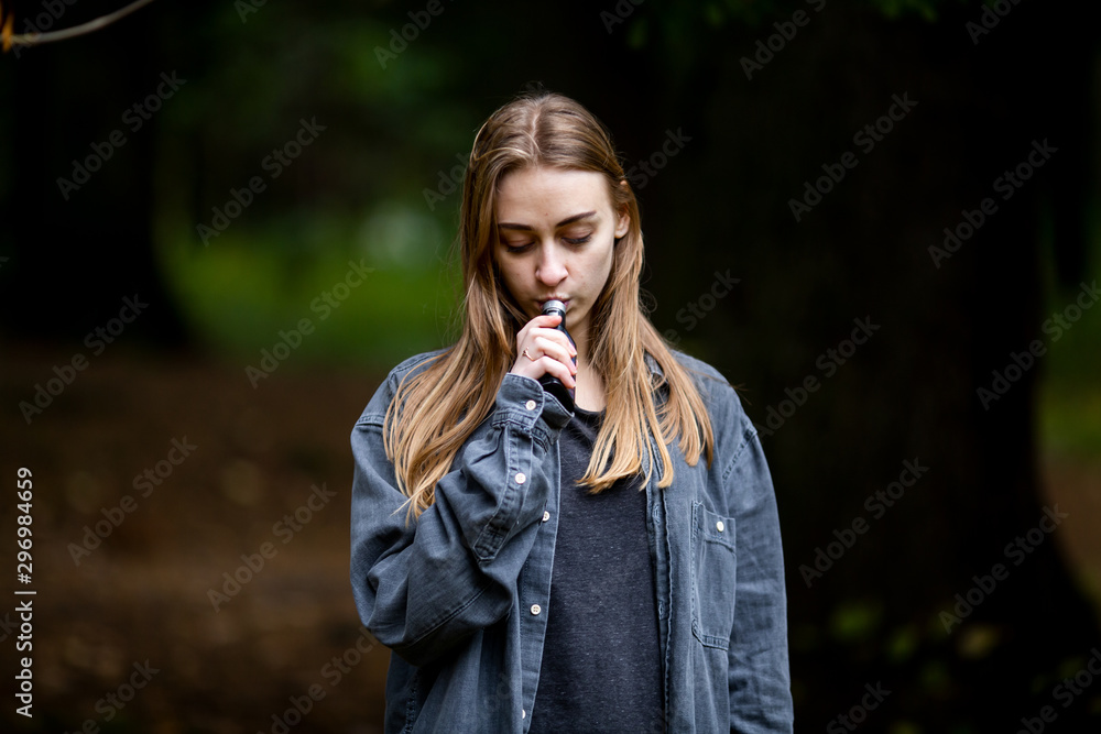 Vape teenager. Young beautiful white teenage girl in casual clothes vaping electronic cigarette on the street in the dark forest in the autumn evening. Bad habit. Vaping activity. Vape teenager. Young beautiful white teenage girl in casual clothes vaping electronic cigarette on the street in the dark forest in the autumn evening. Bad habit. Vaping activity.