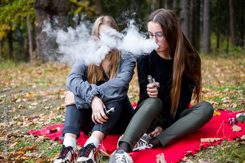 Vape lgbt teenagers. Bisexual lesbian young caucasian teenage girls in casual clothes vape electronic cigarette on the street in the park in the autumn evening. Bad habit.